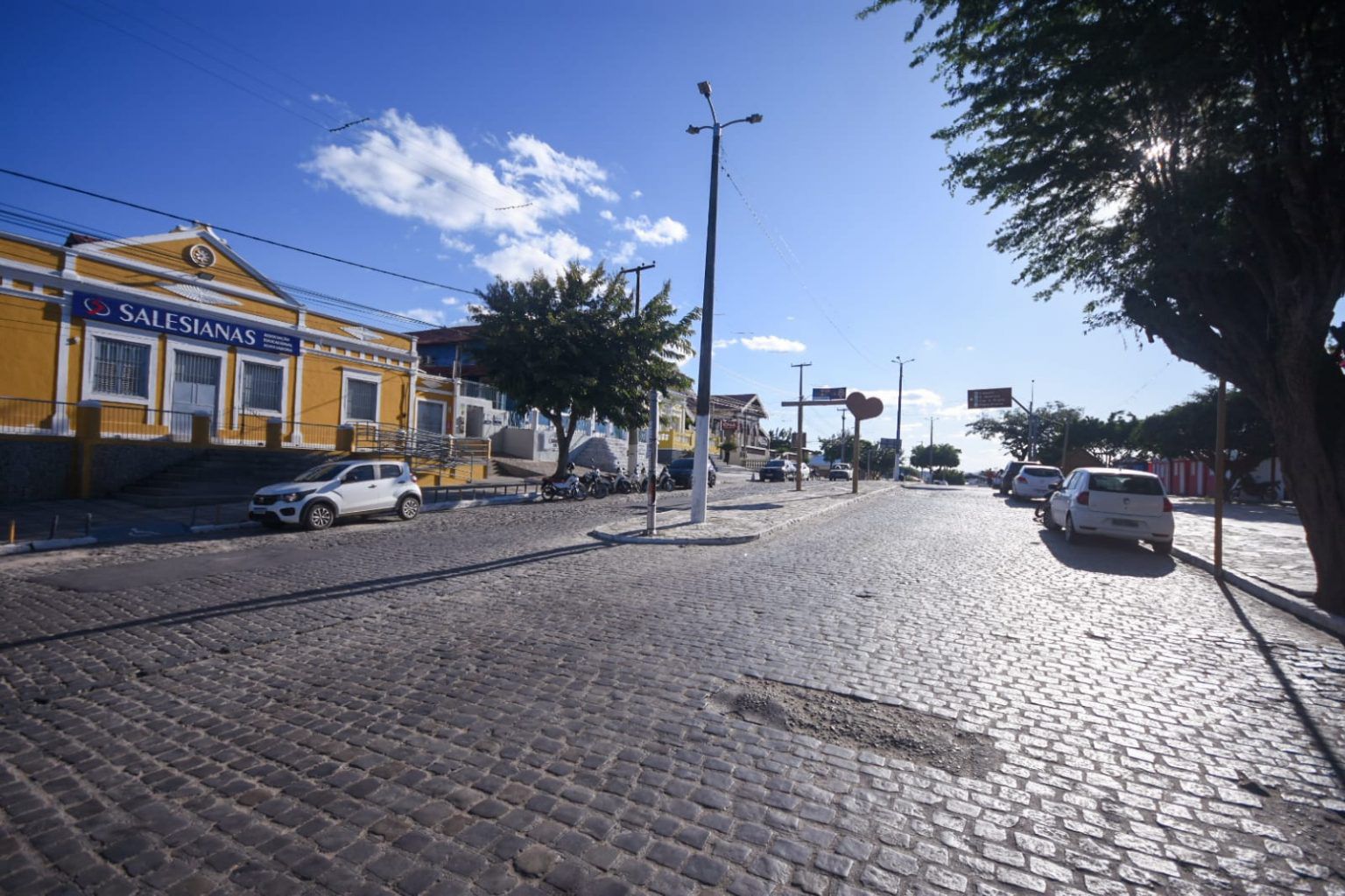 Rua calçada em paralelepípedos em Gravatá, com movimento de carros estacionados e em circulação. À esquerda, destaca-se o prédio amarelo das Salesianas, enquanto à direita há árvores que projetam sombra sobre a via em um dia ensolarado de céu azul.