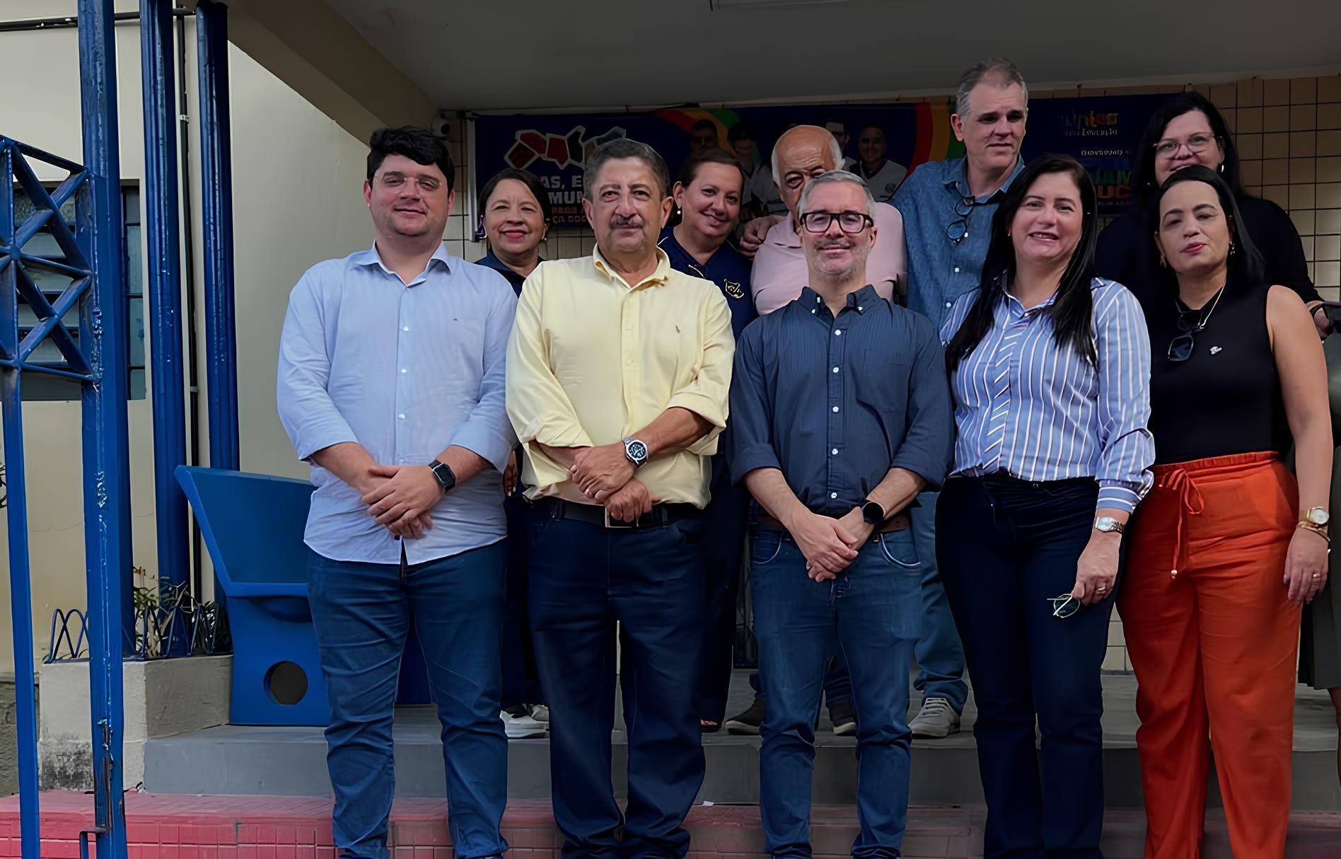 Grupo de homens e mulheres, incluindo o ex-prefeito de Gravatá Joaquim Neto, e o secretário de Educação de Pernambuco, Gilson Monteiro Filho, posam para foto em frente a um prédio institucional, provavelmente após visita ou evento oficial. As pessoas estão sorrindo e organizadas em duas fileiras, com alguns vestindo roupas sociais. Ao fundo, há um banner com informações parcialmente visíveis e paredes revestidas de azulejo claro.