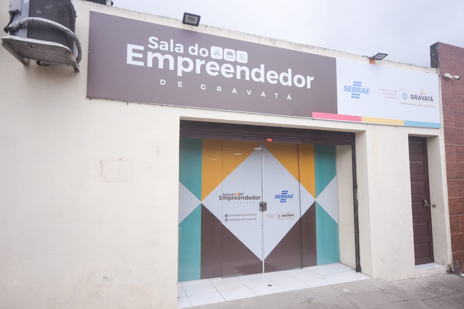 Fachada da Sala do Empreendedor de Gravatá, identificada com placas nas cores marrom e branco, logomarcas do SEBRAE e da Prefeitura Municipal, além de portas de vidro com adesivos coloridos.