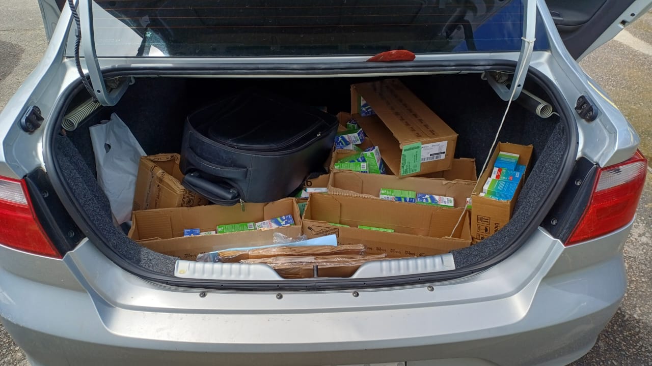 Porta-malas aberto de um carro prata contendo caixas de papelão com diversas embalagens de medicamentos, além de uma mala preta e outros objetos soltos.