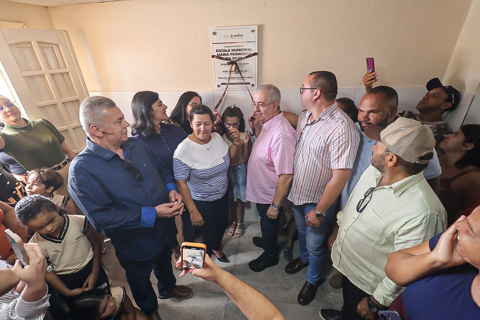Grupo de pessoas participa da cerimônia de reinauguração da Escola Municipal Maria Francisca da Silva, em Gravatá. A placa de requalificação está sendo descerrada por autoridades locais, enquanto crianças, servidores e populares acompanham e registram o momento com celulares.3/3  Perguntar ao ChatGPT
