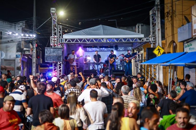 "Apresentação musical ao ar livre durante a noite. Uma banda toca sob uma cobertura branca, com luzes coloridas iluminando o palco.
