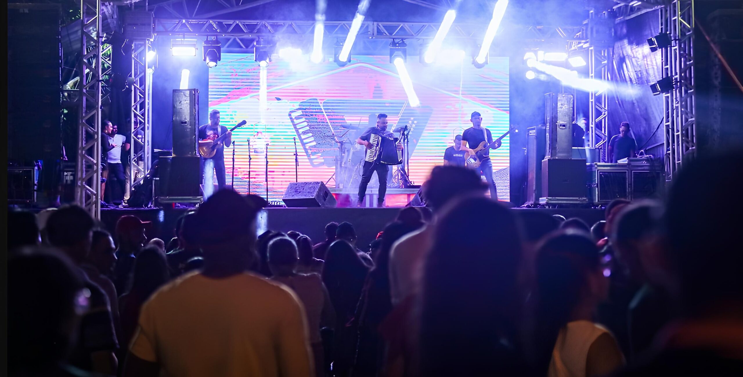 Show ao vivo com banda no palco iluminado, músicos tocando instrumentos como guitarra e sanfona, tela de LED exibindo gráficos coloridos, público assistindo animado em primeiro plano.