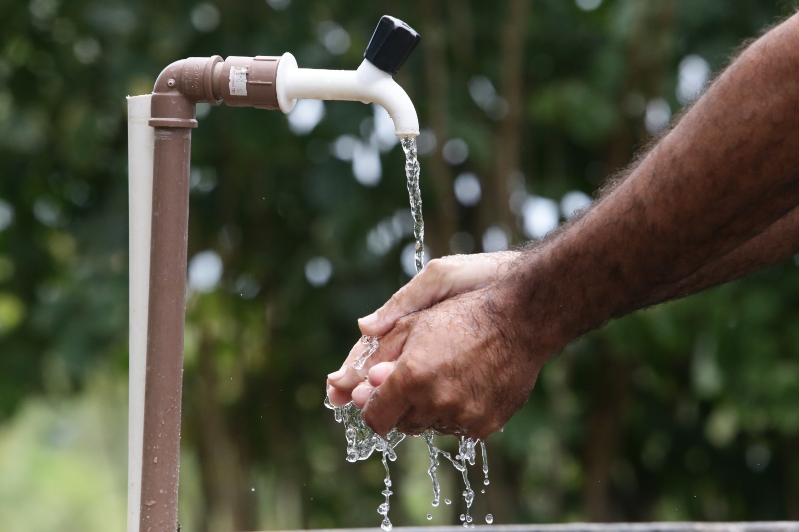 Pessoa lavando as mãos sob uma torneira externa com água corrente, em ambiente natural com folhagem verde ao fundo.