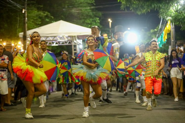Desfile festivo com dançarinos de frevo segurando guarda-chuvas multicoloridos diante de público.