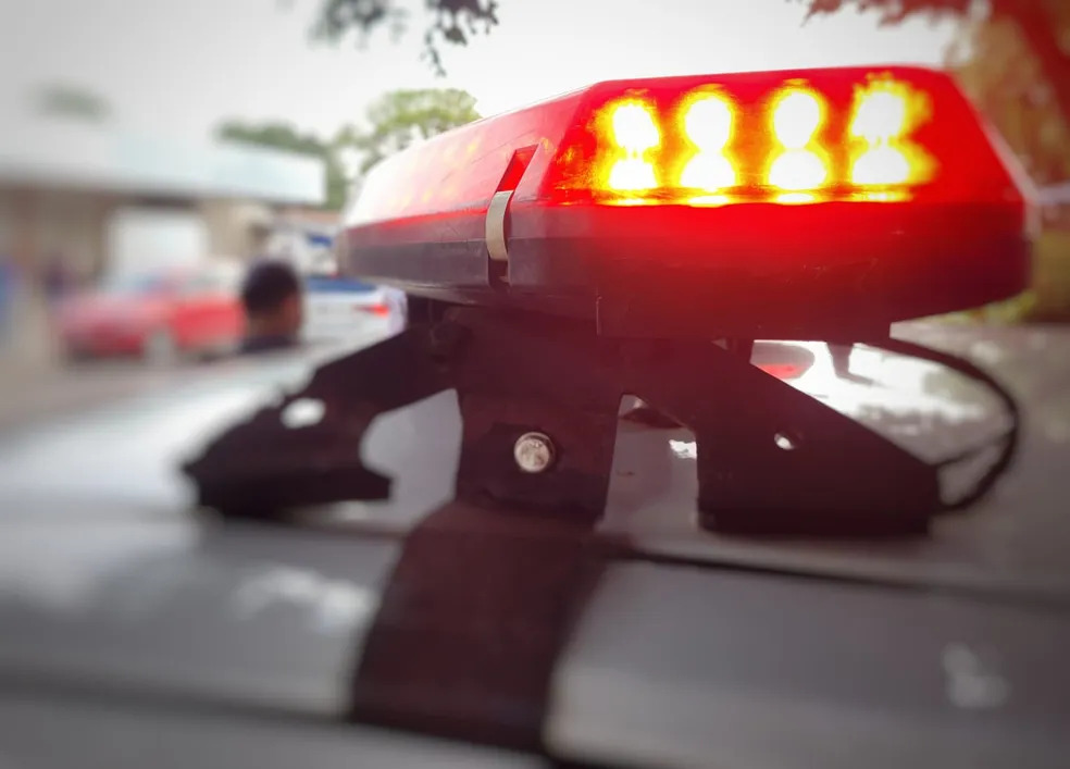 Barra de luz vermelha de emergência acesa no teto de veículo policial, com fundo desfocado mostrando carros e uma pessoa.