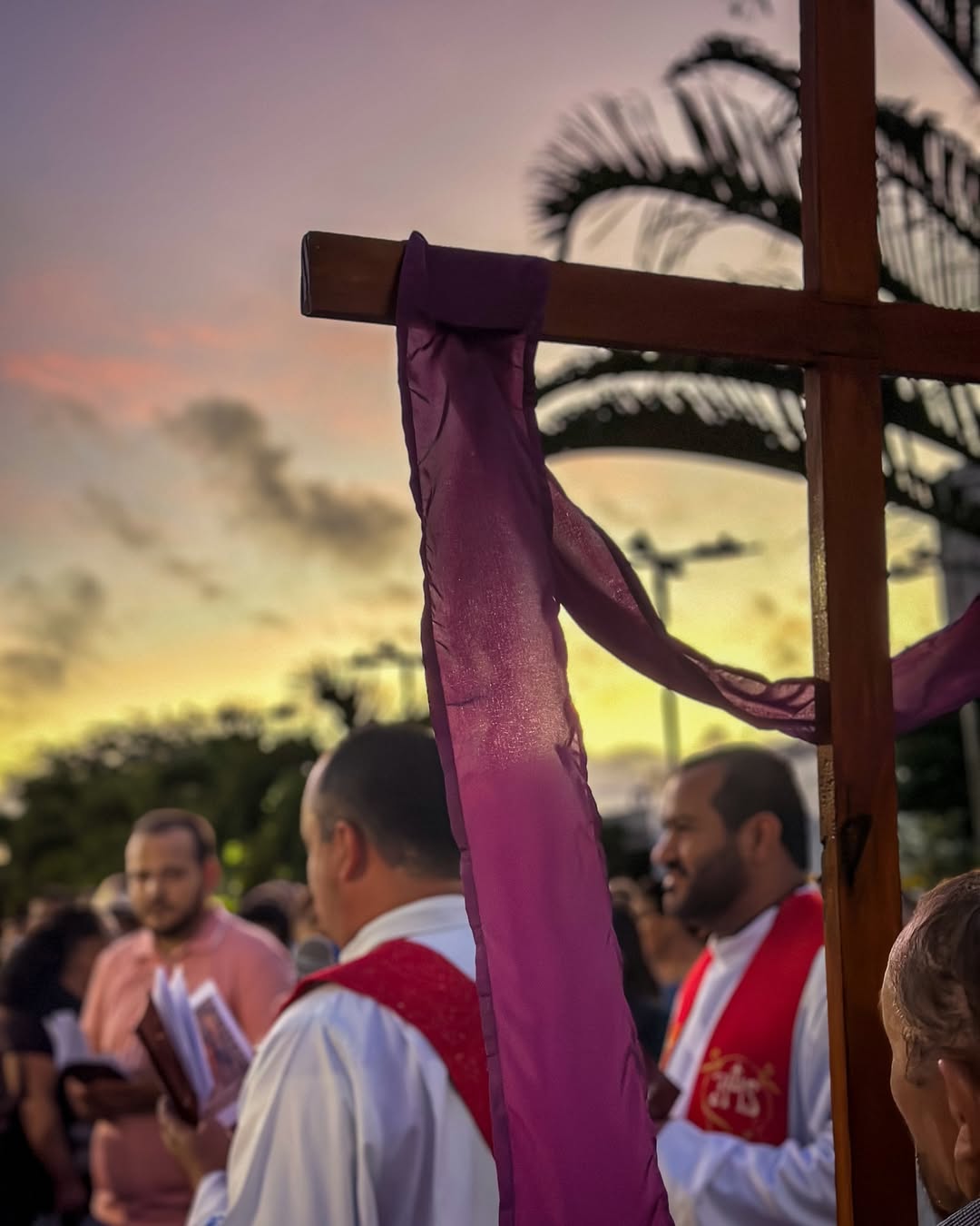 Cruz de madeira com tecido roxo, em frente a pessoas de túnicas brancas e estolas vermelhas durante cerimônia ao pôr do sol.
