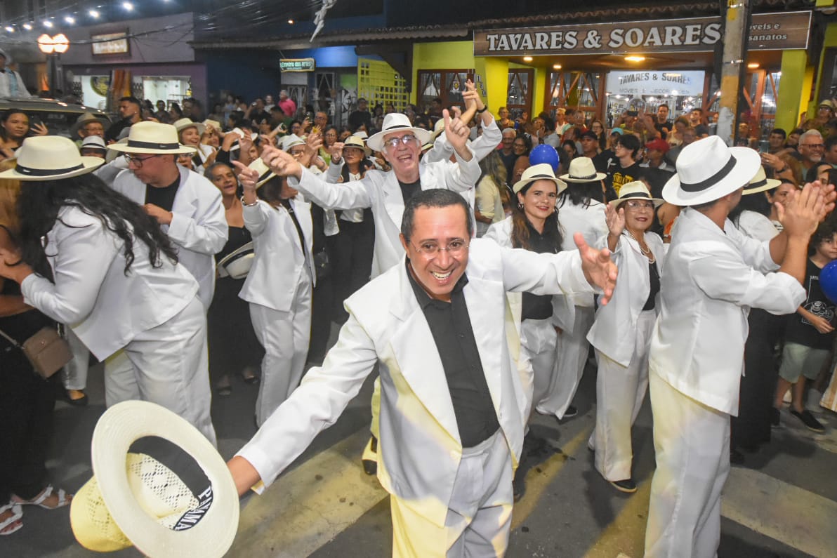 Desfile de Seresteiros em rua iluminada, com grupo de pessoas em trajes brancos e chapéus dançando diante de espectadores.