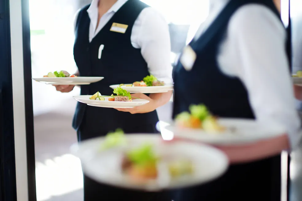 Dois garçons em uniforme formal segurando pratos de comida em ambiente de restaurante ou evento.