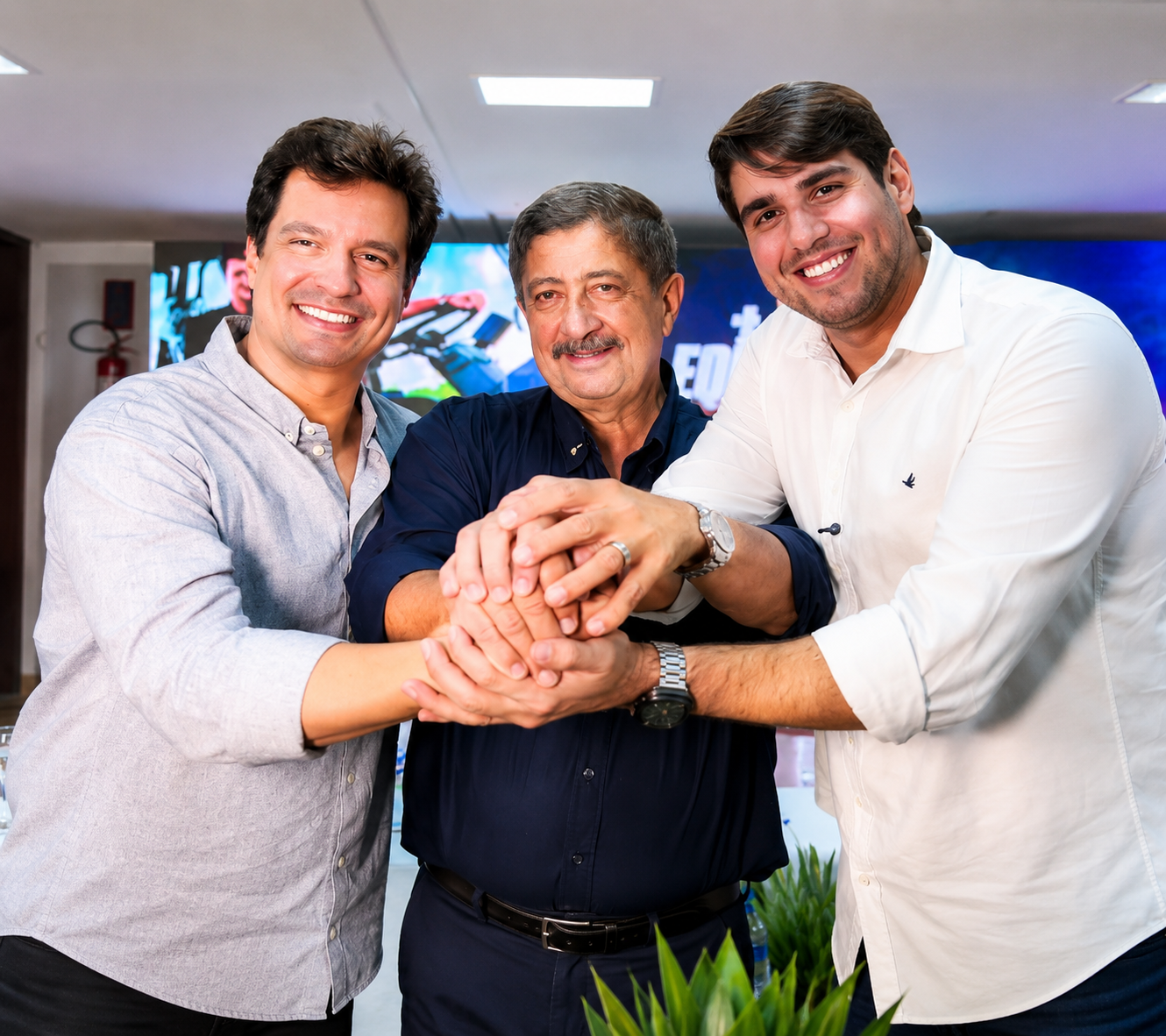 Joaquim Neto, ao centro, ladeado por Lucas Ramos (à esquerda) e Bruno Marques (à direita), posa sorrindo em evento político, com as mãos unidas ao centro, em ambiente interno com painel iluminado ao fundo.