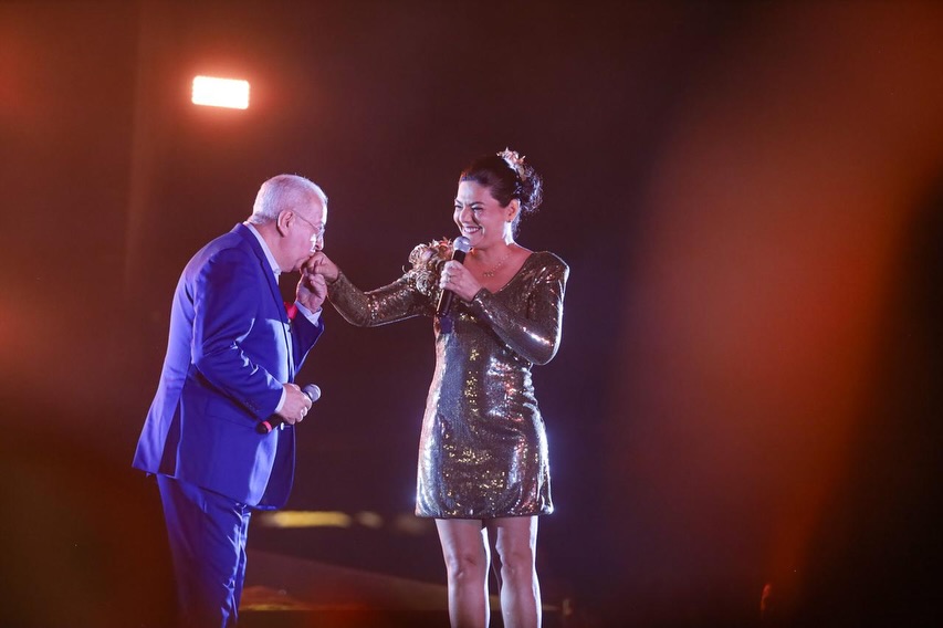 Prefeito Joselito Gomes, de terno azul, segura um microfone e beija a mão da primeira-dama Viviane Facundes durante apresentação em palco iluminado. Viviane, vestindo um traje brilhante e também com microfone na mão, sorri para ele sob luzes alaranjadas ao fundo.