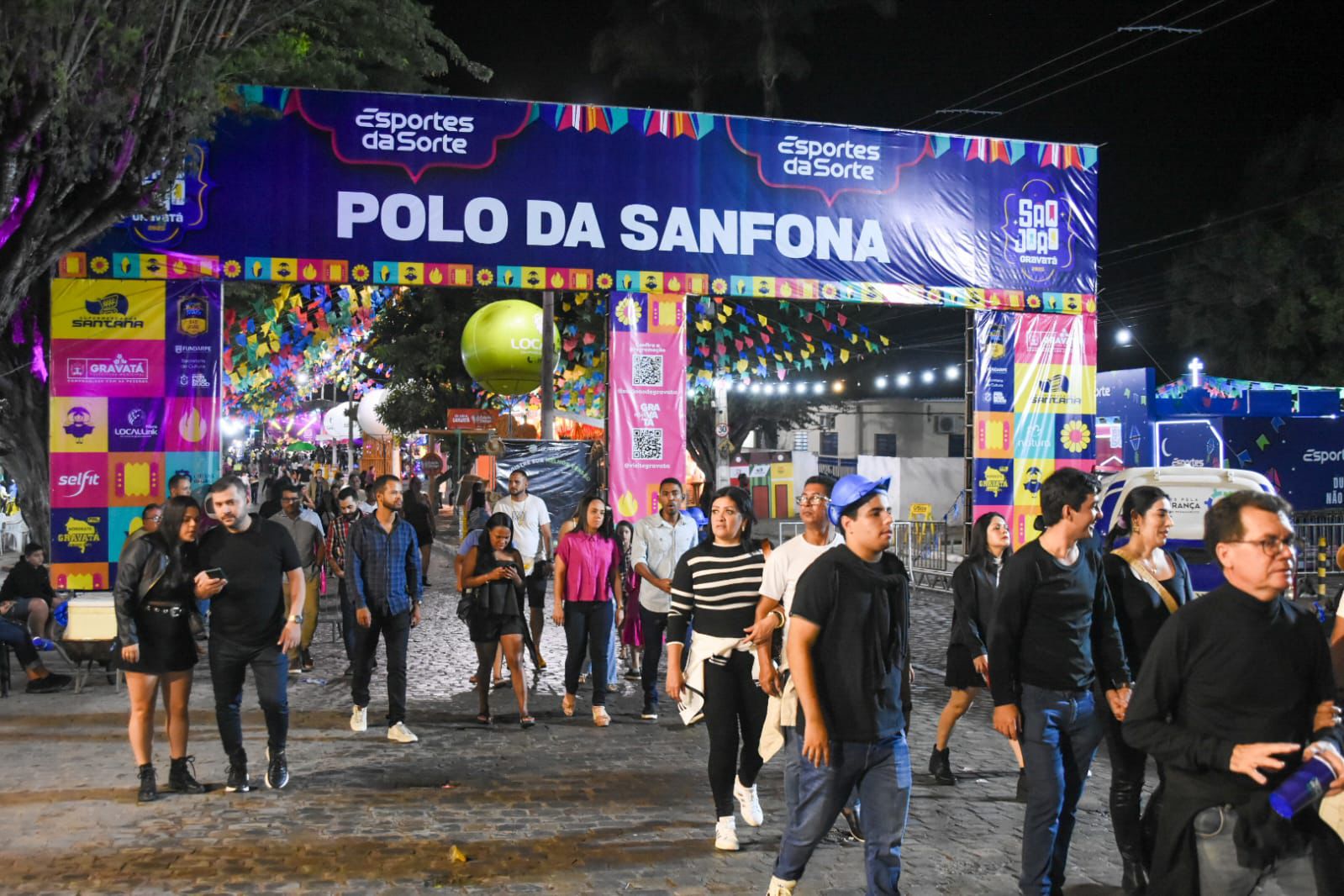 Arco iluminado escrito ‘Polo da Sanfona’, com bandeirinhas coloridas e pessoas caminhando em rua de paralelepípedos à noite.